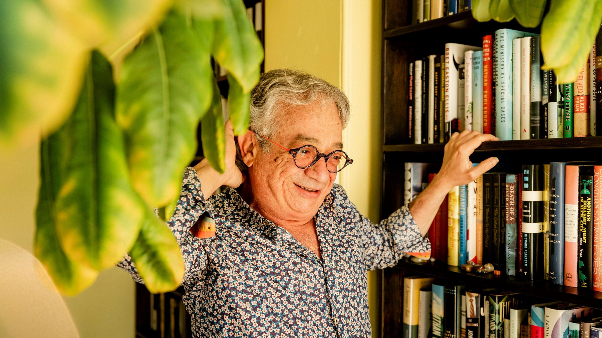 Rabih Alameddine stands between a bookshelf and a plant. He wears a bright floral patterned shirt and round black and red speckled glasses.