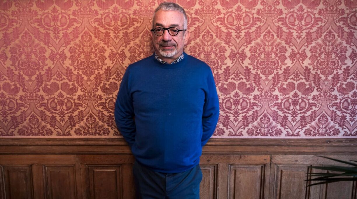 Headshot of Rabih Alameddine in front of a wall with pink floral wallpaper. Alameddine is wearing a royal blue sweater and blue slacks.