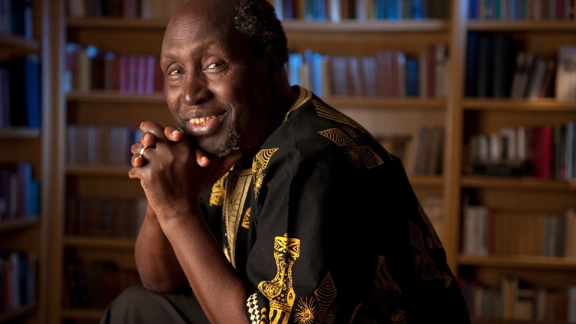 Ngũgĩ wa Thiong'o smiling with arms resting on his chin, in black and gold shirt.