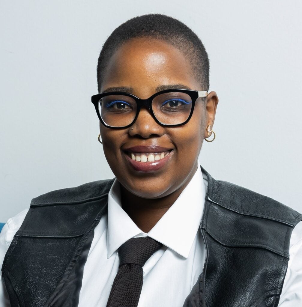 Novuyo Rosa Tshuma with short hair and wearing black square rimmed glasses, white button down with black leather vest, and black tie.