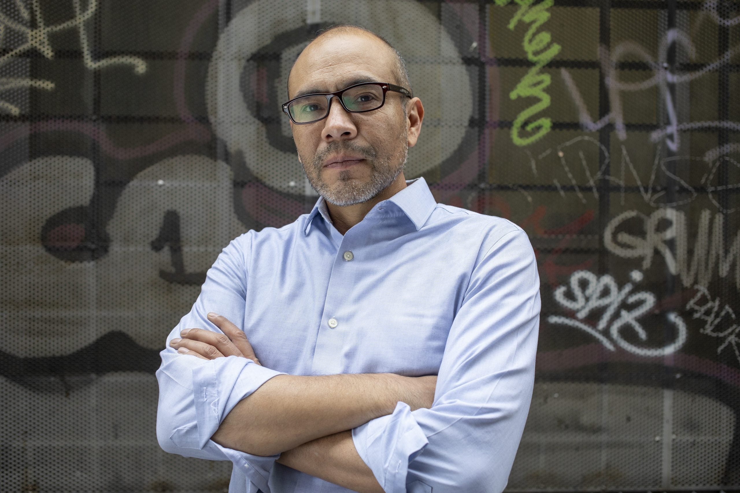 Yuri Herrera in a light blue dress shirt and black rimmed glasses, with arms crossed in front of a graffiti wall.