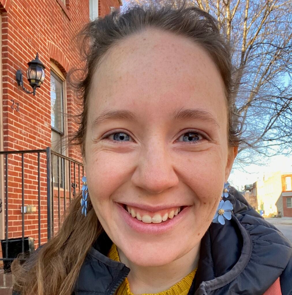 A white female smiling, wearing her hair in a ponytail and blue flower earrings, with a red brick row home and winter tree in the background.