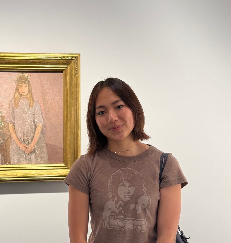 An Asian female with short brown hair wears a brown Doors t-shirt and is standing in front of a gold-framed painting of a young girl at a gallery.