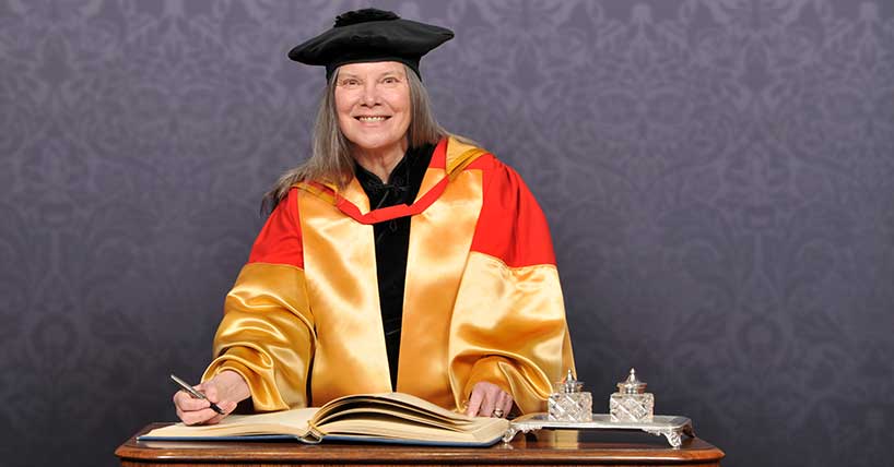 Carolyn Forché is dressed in yellow and orange doctoral robes. She is smiling at the camera as she is signing a book of records.