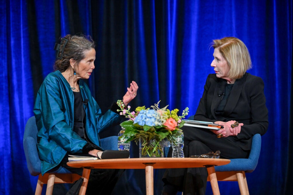 Two people are sitting down in a conversation. Behind them is a blue satin curtain, and front of them is a low table with a small vase of flowers. Julia Alvarez is seated on the left and is wearing a blue cardigan over a black shirt. Georgina Godwin is seated on the right and is wearing a full black formal feminine suit.