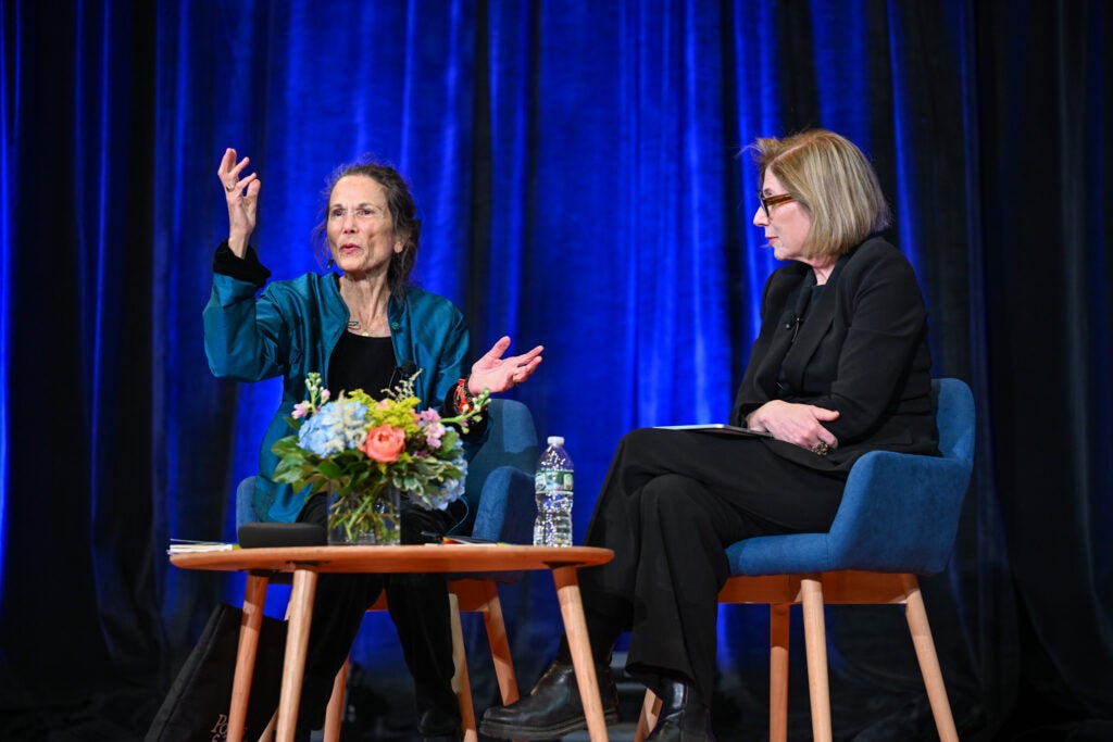 Two people are sitting down in a conversation. Behind them is a blue satin curtain, and front of them is a low table with a small vase of flowers. Julia Alvarez is seated on the left and is wearing a blue cardigan over a black shirt. Georgina Godwin is seated on the right and is wearing a full black formal feminine suit.