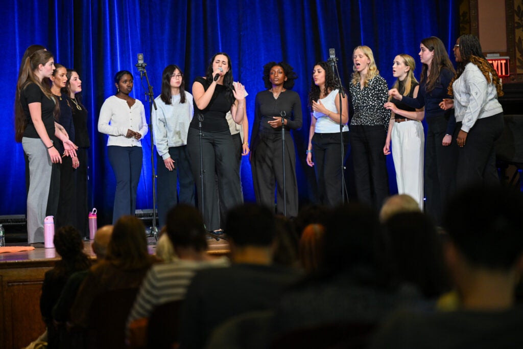 The GraceNotes is singing a performance on a stage. Behind them is a blue satin curtain. A seated crowd is watching.