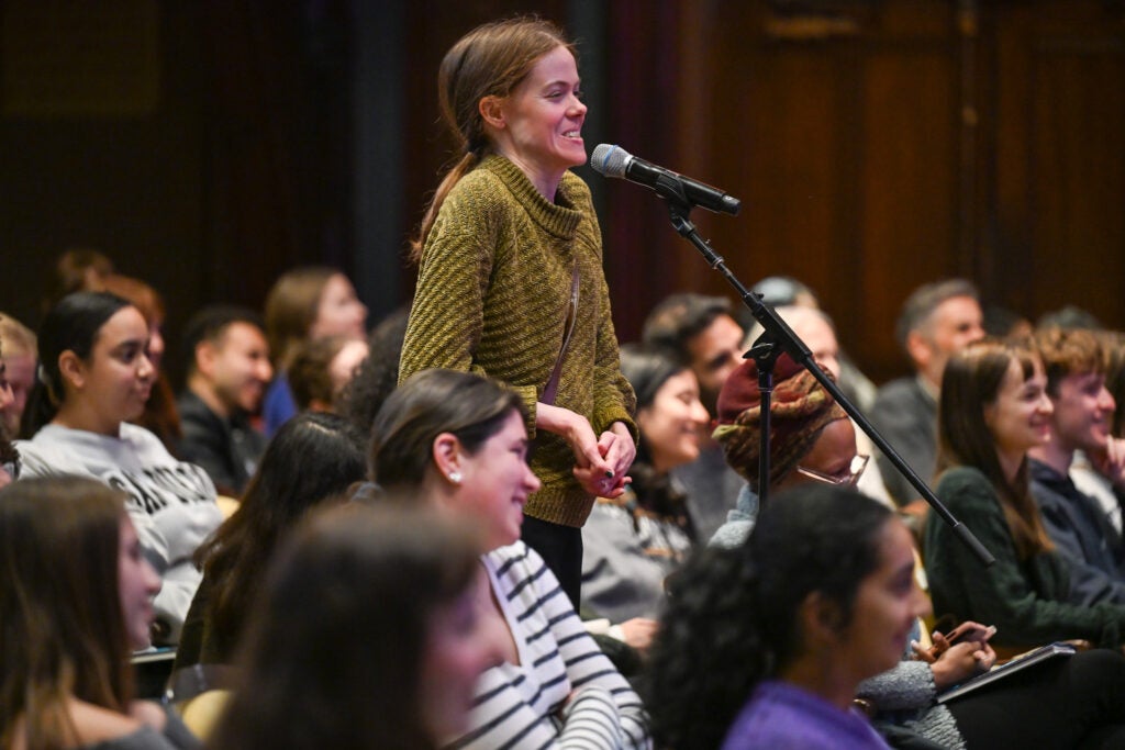 A person in a green sweater is speaking to a microphone stand. Around the speaker in green is a seated crowd who are laughing.