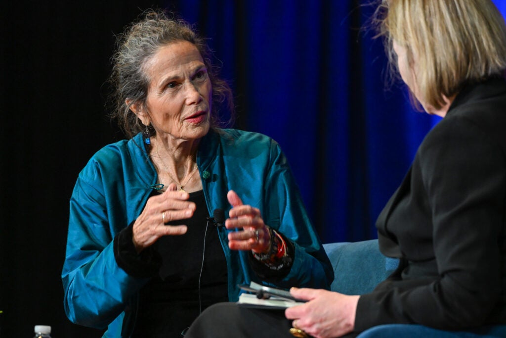 Julia Alvarez is seated on the left and is wearing a blue cardigan over a black shirt. Julia is speaking to Georgina. Georgina Godwin is seated on the right and is wearing a full black formal feminine suit.